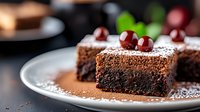 Rich chocolate brownie slice dusted with cocoa powder and topped with fresh cherries on white plate, selective focus with blurred background. Appetizing dessert.