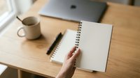Woman's hand opening spiral notebook on wooden desk with laptop, pen and coffee mug for work, study, planning and creative projects.