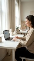 Young professional woman working on laptop at home office desk by window, smiling while typing. Remote work, freelancing, and modern workspace concept.