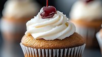Fresh baked vanilla cupcake topped with swirled white buttercream frosting and garnished with bright red maraschino cherry against dark background.