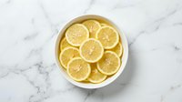 Fresh lemon slices arranged in white bowl on marble surface. Citrus fruit preparation for cooking, beverages, garnish and healthy nutrition concepts.