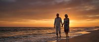 Romantic couple holding hands walking on beach at golden sunset with dramatic orange sky and ocean waves creating peaceful moment for love and togetherness.