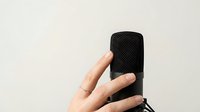 Hand holding black wireless portable speaker with mesh grille against neutral beige background, isolated for design use in technology and audio concepts.