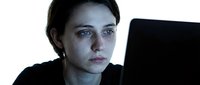 Young Caucasian woman with tired expression working late on laptop computer in dark room with screen glow illuminating face showing exhaustion and stress.