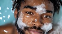 Young African American man with facial foam cleanser smiling during skincare routine against blurred bathroom lights background. Wellness and self-care concept.
