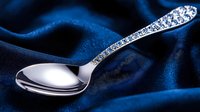 Silver spoon with ornate handle pattern on dark blue silk fabric background, elegant tableware detail in dramatic lighting for luxury dining and decoration.
