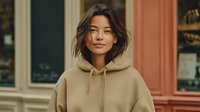 Young Asian woman in beige hoodie standing confidently outdoors near colorful building facade for casual fashion and lifestyle content.