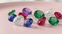 Collection of colorful precious gemstones including diamonds, emeralds, sapphires and rubies scattered on soft pink surface for luxury jewelry design.