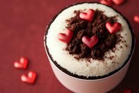 Romantic dessert with chocolate crumbs and pink heart decorations on creamy white topping in ceramic cup against red background, selective focus macro shot.