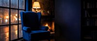 Cozy reading nook with blue velvet armchair, warm lamp, fireplace glow, and bookshelf in moody evening ambiance. Perfect for interior design and lifestyle concepts.