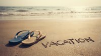 Flip flops on sunny beach sand with vacation word written nearby, ocean waves in background, warm summer holiday travel concept.