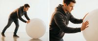 Young athletic man in black hoodie pushing large white exercise ball during fitness workout training session in modern gym studio.