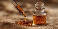 Dropper dispensing golden amber essential oil into small glass bottle on sandy textured background.