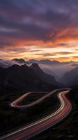 Dramatic mountain highway with light trails at sunset showcasing winding road through alpine landscape with purple orange sky and layered peaks in distance.