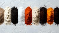 Colorful spices and seasonings arranged in neat rows on white textured background, including paprika, black pepper, turmeric, salt and other ground powders.