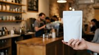 Barista holding blank white pouch in coffee shop with customers and counter in background, suitable for packaging mockups and cafe branding.