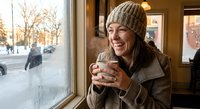 Young Asian woman in knit beanie enjoying hot beverage by cafe window during winter, smiling warmly while looking outside at snowy street scene.