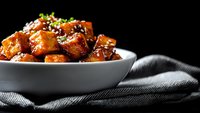 Glazed chicken pieces garnished with sesame seeds and green onions in white ceramic bowl against dark background, dramatic food photography style.