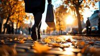 Person walking on autumn street with shopping bag during golden sunset, fallen orange leaves scattered on sidewalk, creating atmospheric urban scene.