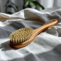 Natural wooden body brush with soft bristles on white cotton fabric background, creating gentle shadows in soft daylight. Eco-friendly spa and wellness concept.