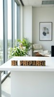 Wooden letter blocks spelling "BE PREPARED" on white desk in bright modern office with large windows, green plant, and minimalist decor for motivational business concepts.