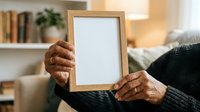 Senior woman with dark skin holding wooden frame with blank white space, wearing black sweater in cozy home interior with shelves and warm lighting for memorial, memory, or family photo display.
