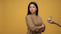 Young Middle Eastern woman with crossed arms looking skeptical at smartphone held by another person against yellow background for social media concepts.