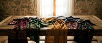 Colorful vintage clothing collection spread across wooden table in rustic stone interior with natural window light for fashion styling concepts.