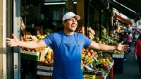 Happy Hispanic male vendor in blue apron welcoming customers at fresh produce market stall with colorful fruits and vegetables display for small business.