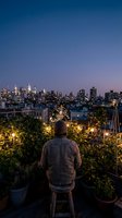 Man sitting on rooftop garden overlooking illuminated city skyline at dusk, urban gardening and contemplation concept for lifestyle and real estate projects.