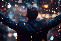 Businessman with raised arms celebrating success against night city lights and bokeh effect, rear view showing achievement and triumph mindset.