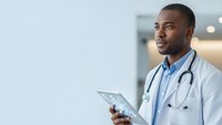 Young African American male doctor in white coat holding tablet computer with stethoscope around neck smiling confidently in modern hospital setting.
