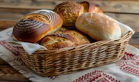 Fresh baked bread rolls and buns with various toppings arranged in wicker basket on rustic wooden table with traditional embroidered cloth, warm natural lighting.