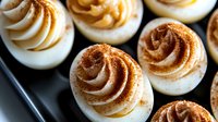 Deviled eggs topped with creamy mustard filling piped into decorative swirls, garnished with paprika powder, arranged on a dark plate for appetizer or party food presentation.
