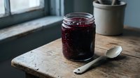 Homemade dark berry jam in glass jar on rustic wooden cutting board with vintage spoon, natural kitchen lighting for breakfast and baking recipes.