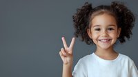 Young African American girl with curly hair in white t-shirt showing peace sign gesture against gray background, smiling with bright cheerful expression.
