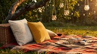 Cozy outdoor picnic setup with wicker basket, decorative pillows, blanket, hanging lanterns, and wine glasses under tree at golden hour for entertaining and leisure.