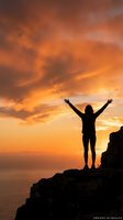 Woman silhouette with raised arms celebrating success on mountain cliff against dramatic orange sunset sky over ocean for motivation concepts.