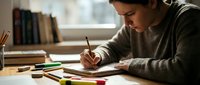 Young woman student writing in notebook at desk with colored pencils and books in bright study room for education and learning projects.