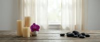 Spa wellness setup with candles, purple orchid, and black zen stones on rustic wooden table near sheer curtained window, soft light.