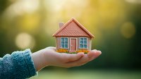 Small pink house model with orange flowers resting in female hand wearing blue sweater against blurred green and golden bokeh background. Concept of real estate and home ownership.