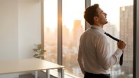 Confident businessman in white shirt adjusting tie while looking out office window at sunset city skyline, corporate success and achievement concept.