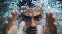 Young man with intense gaze emerging from water with dramatic splash effect, hands raised creating dynamic water movement. Artistic portrait capturing raw emotion and natural elements.