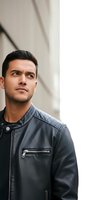 Young man of Middle Eastern descent wearing black leather jacket over denim shirt, looking upward thoughtfully against urban wall background. Suitable for lifestyle, fashion, and portrait projects.