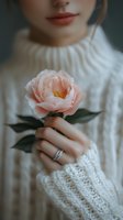Cropped view of woman in white knit sweater holding delicate pink rose, wearing elegant diamond ring. Soft moody atmosphere with focus on feminine details.