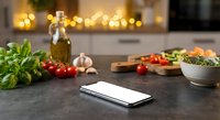 Smartphone on kitchen counter with fresh ingredients, olive oil, herbs and vegetables for cooking recipes and meal planning apps.
