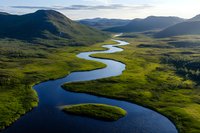 Meandering river curves through green valley surrounded by mountains, aerial view of pristine wilderness landscape with winding waterway and lush meadows.