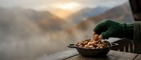 Young person in green sweater holding bowl of nuts outdoors with misty mountain landscape background, autumn season, cozy lifestyle moment.