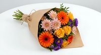 Colorful mixed flower bouquet wrapped in brown kraft paper with orange gerberas, yellow roses, pink chrysanthemums and purple blooms on white background.