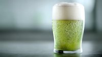 Frosty glass of green beer with foam head and condensation droplets on blurred background, suitable for St Patrick's Day celebrations and bar promotions.